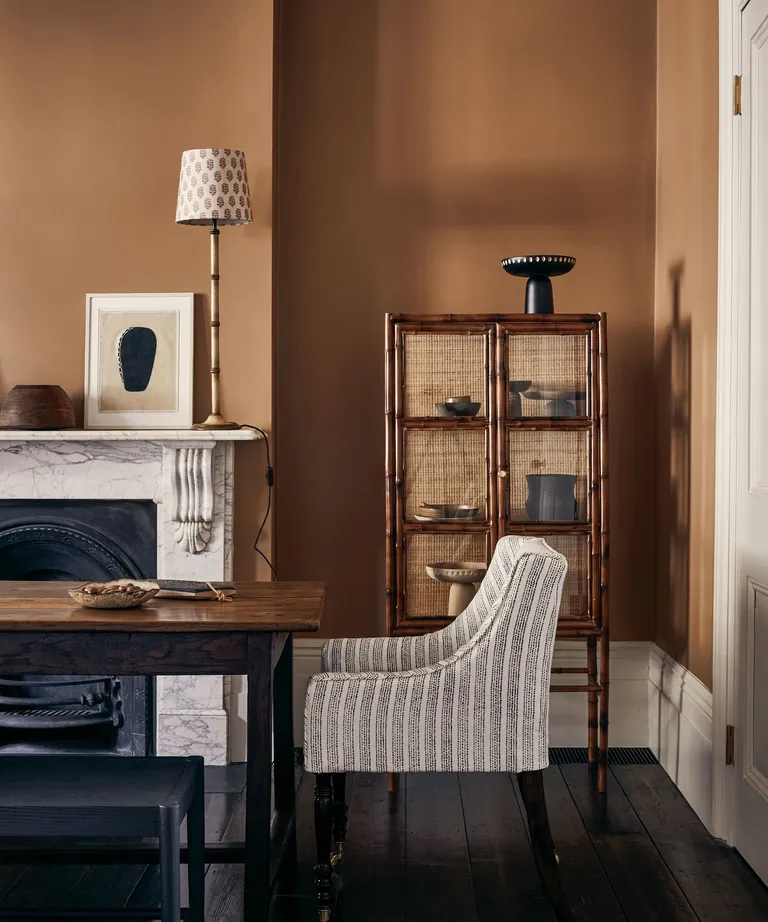 Painted dining room in a dark ochre shade, natural textures and colors used throughout, dark wooden storage cabinet with rattan doors, dark wooden rectangular dining table, striped upholstered dining chair, dining bench, marble fireplace with table lamp, black painted wooden flooring