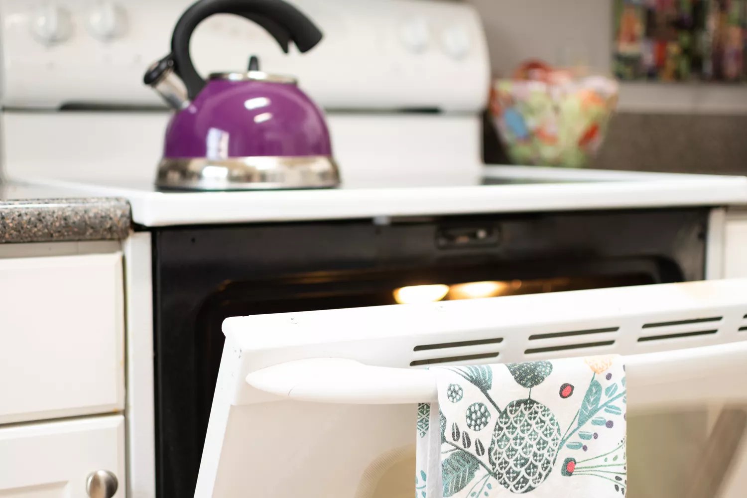 Oven door open with patterned towel hanging and purple teapot on top of stove